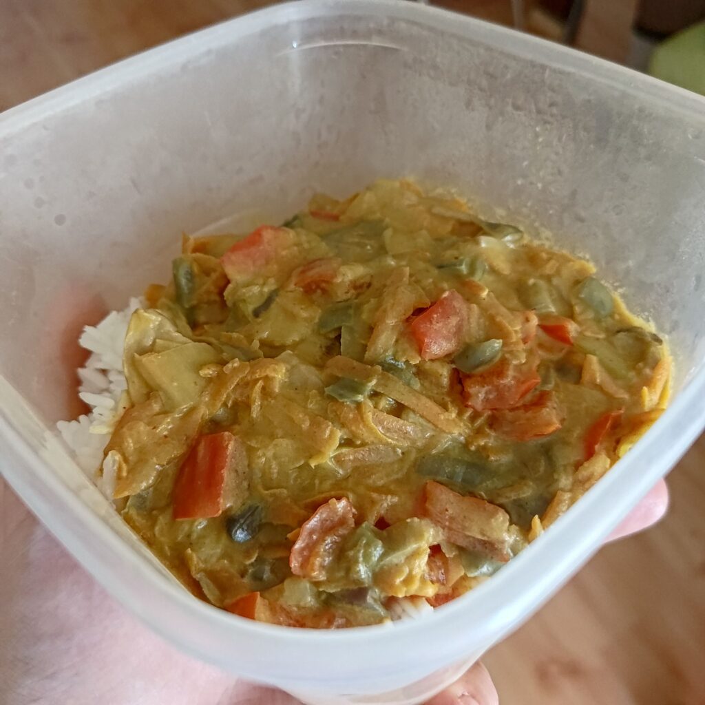 Zdjęcie przedstawiające plastikowy pojemnik z curry z warzywami podanym na ugotowanym białym ryżu
