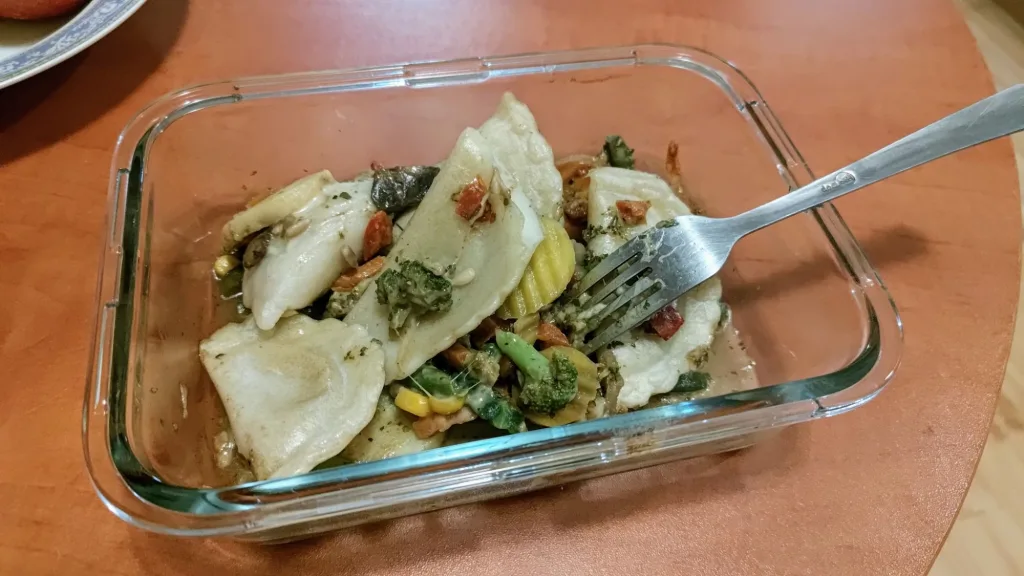 Szklany pojemnik z pierogami, serem mozarella i mieszanką warzyw (brokuły, kukurydza, marchew), jedzone widelcem.