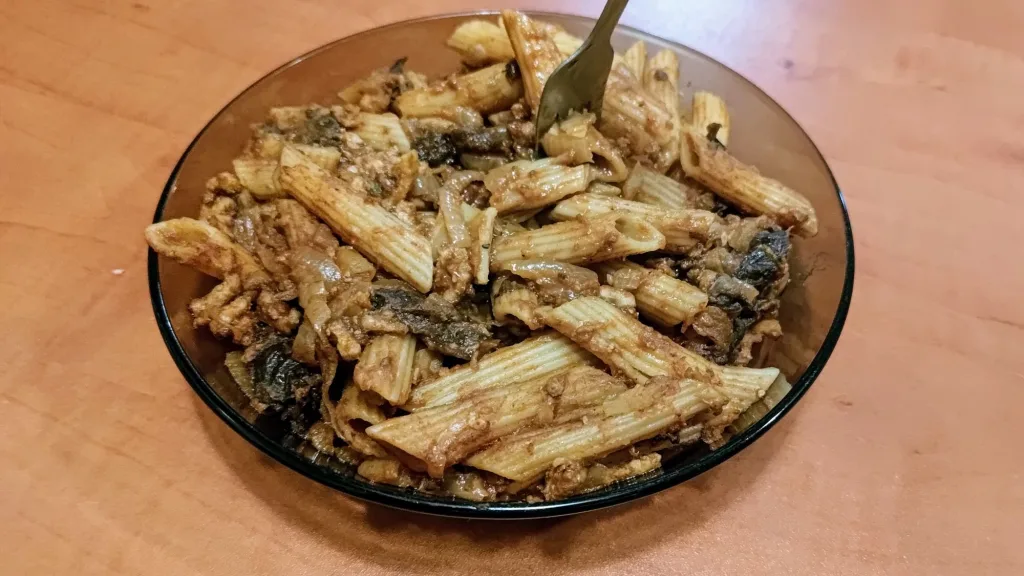 Makaron penne w ciemnym sosie barbecue z widocznymi kawałkami cebuli lub śliwek, z widelcem na głębokim talerzu.
