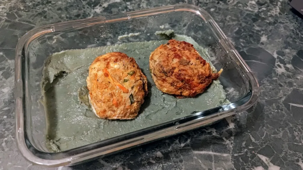 Szklany pojemnik z jedzeniem zawierający dwa usmażone kotlety i zielone puree.