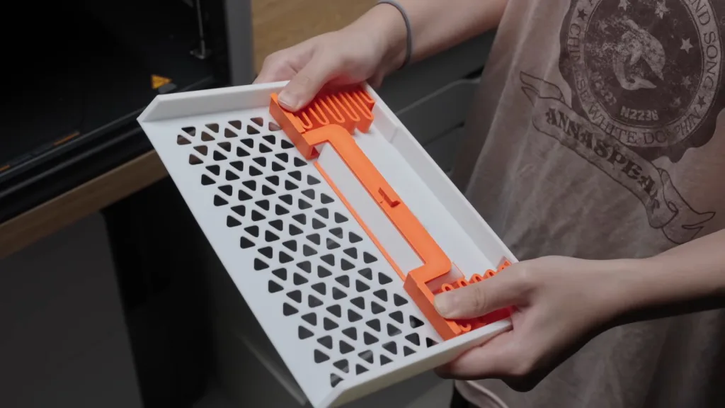 Dłoń trzyma białą tackę z pomarańczową plastikową sprężyną, z otworami w kształcie trójkątów. Jest to element konsoli Ningtendo PXbox 5