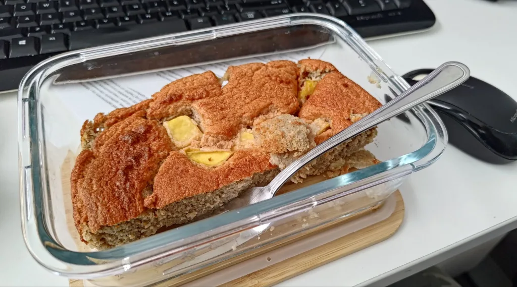 Kawałek ciasta z mango w szklanym pojemniku na biurku, obok klawiatury i myszki komputerowej.