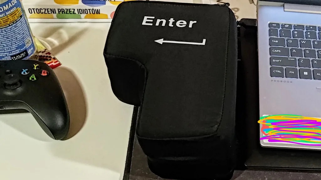 Czarny, miękki pokrowiec w kształcie klawisza Enter z białym napisem "Enter" i ikoną strzałki, obok znajduje się kontroler do gier i laptop.