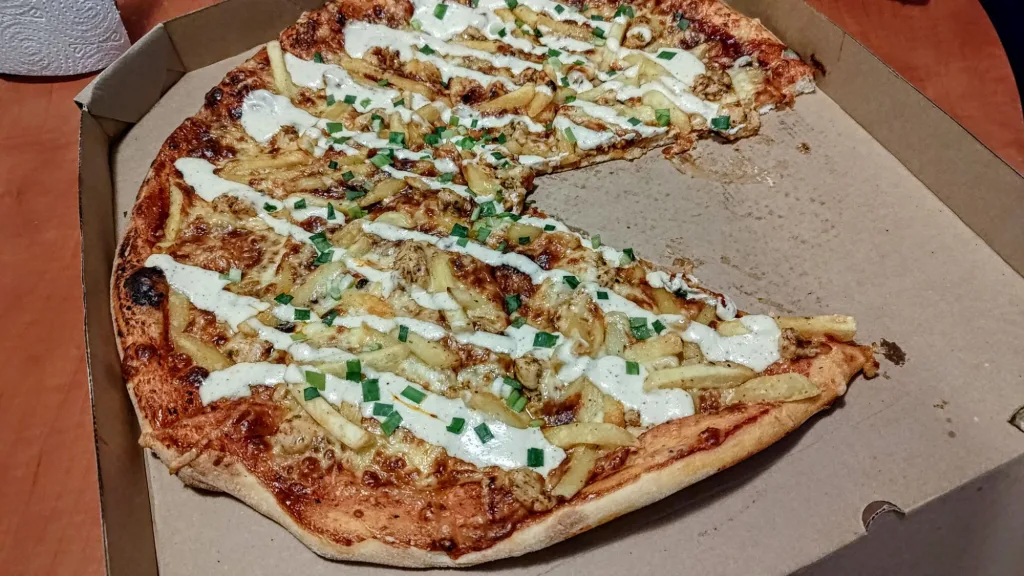 Pizza z frytkami, kurczakiem i sosem na cieście z serem, posypana szczypiorkiem, w kartonowym pudełku.