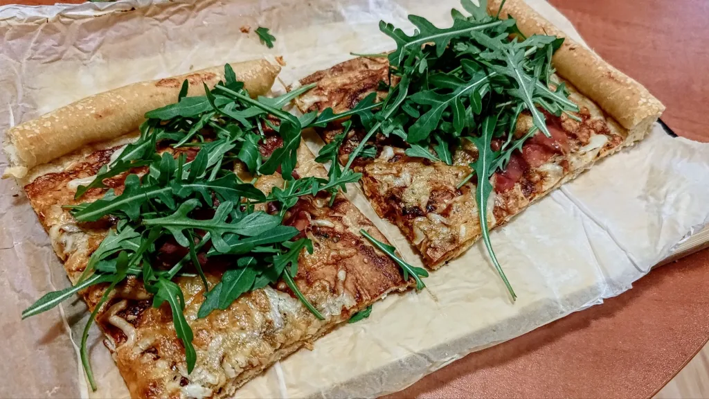 Prostokątna pizza z rukolą i serem, podana na papierze do pieczenia.