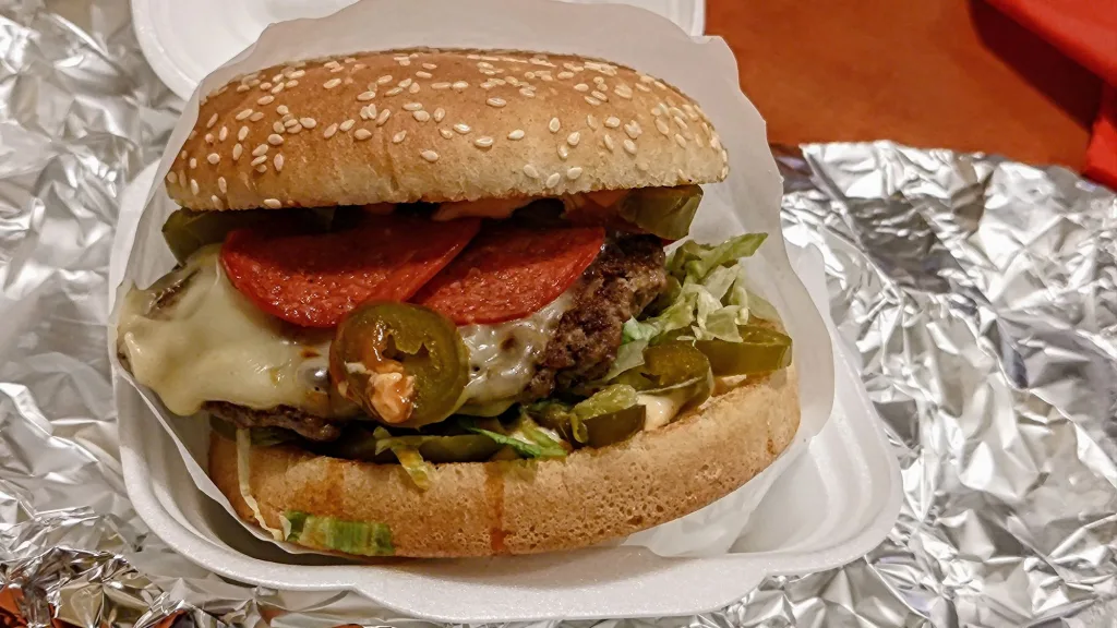 Burger z serem, plasterkami pepperoni i plasterkami jalapeno w bułce z sezamem, podany w papierowym opakowaniu na tle folii aluminiowej.