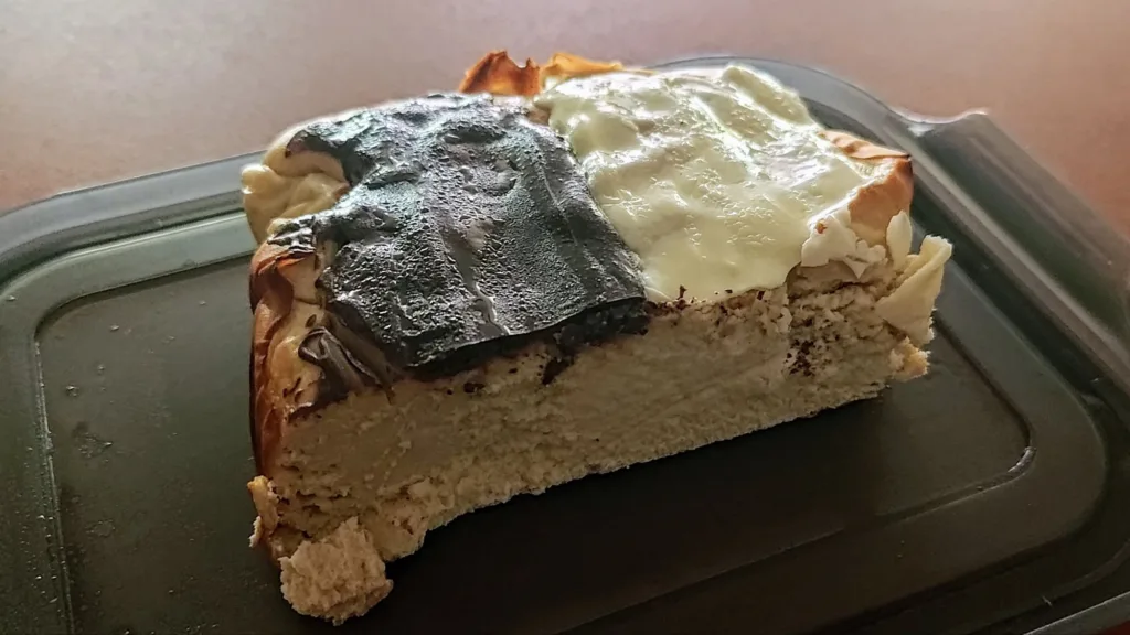 Kawałek sernika z przypaloną górą i białą polewą na ciemnej tacy.