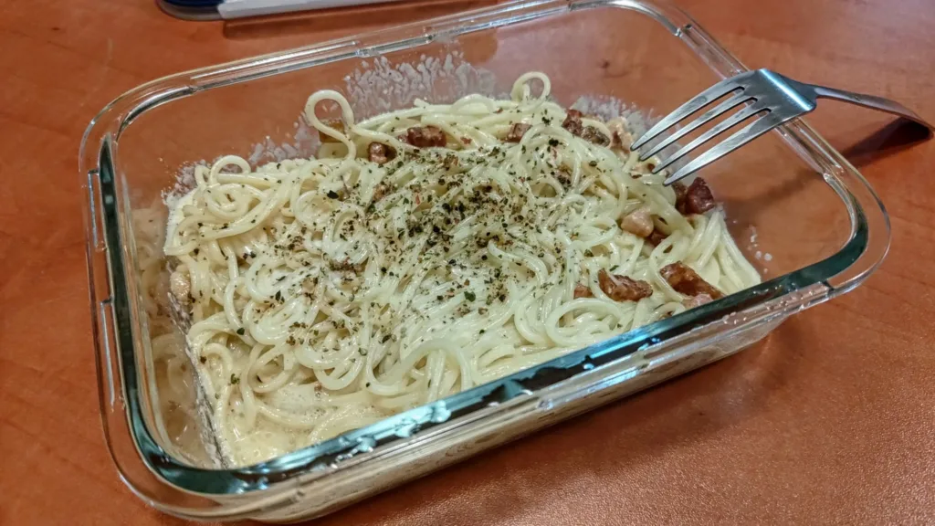 Porcja makaronu spaghetti w sosie śmietanowym z kawałkami bekonu i posypką z ziół, podana w szklanym pojemniku.