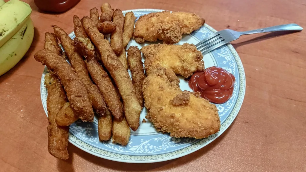 Talerz z frytkami, kawałkami panierowanego kurczaka i ketchupem.