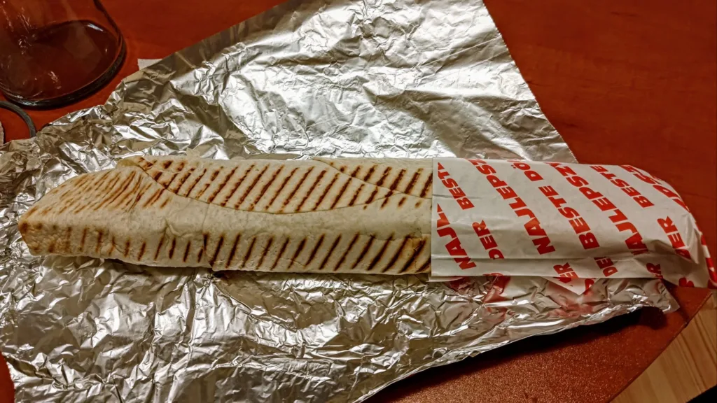 Döner kebab zawinięty w papier z nadrukiem "NAJLEPSZY DER" na tle folii aluminiowej i drewnianego stołu.