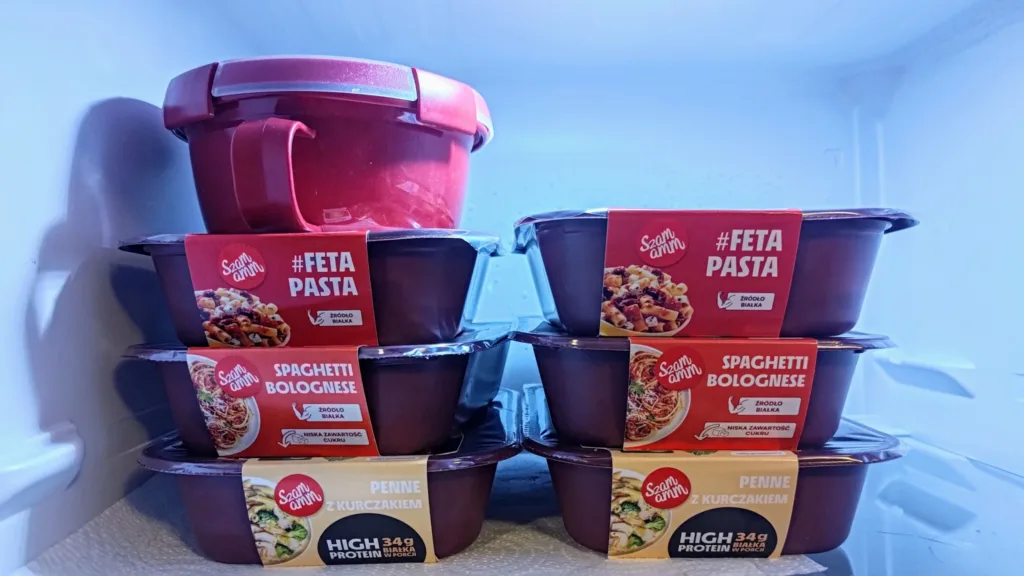 Stos gotowych dań w opakowaniach, w tym #Feta Pasta, Spaghetti Bolognese i Penne z kurczakiem, ustawionych w lodówce.