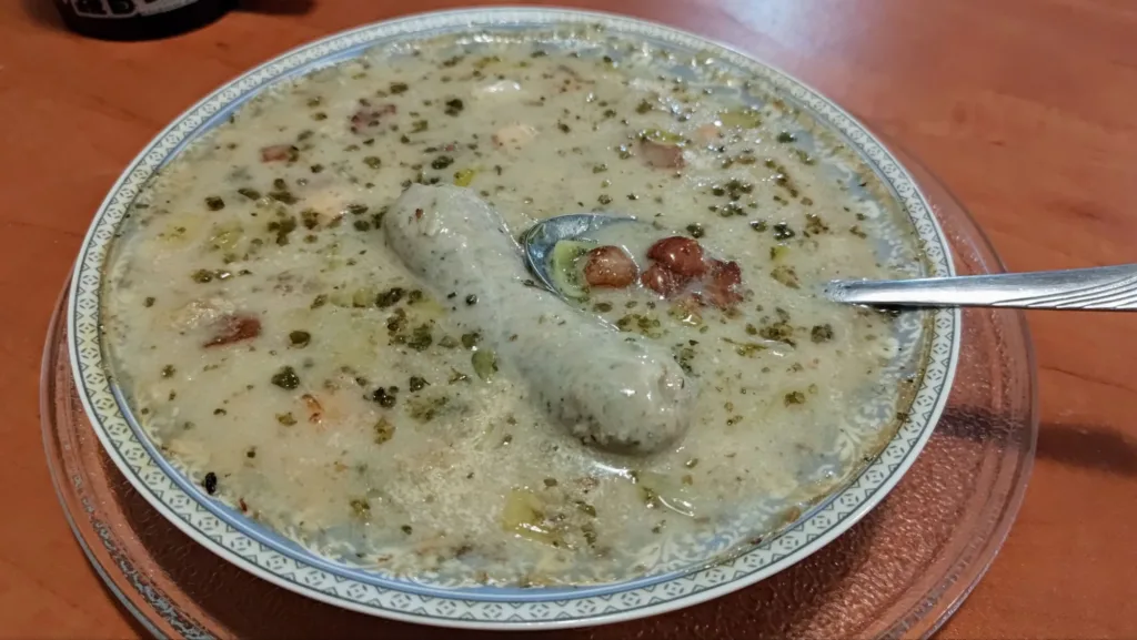 Talerz z zupą, w której znajduje się duża biała kiełbasa i łyżka.