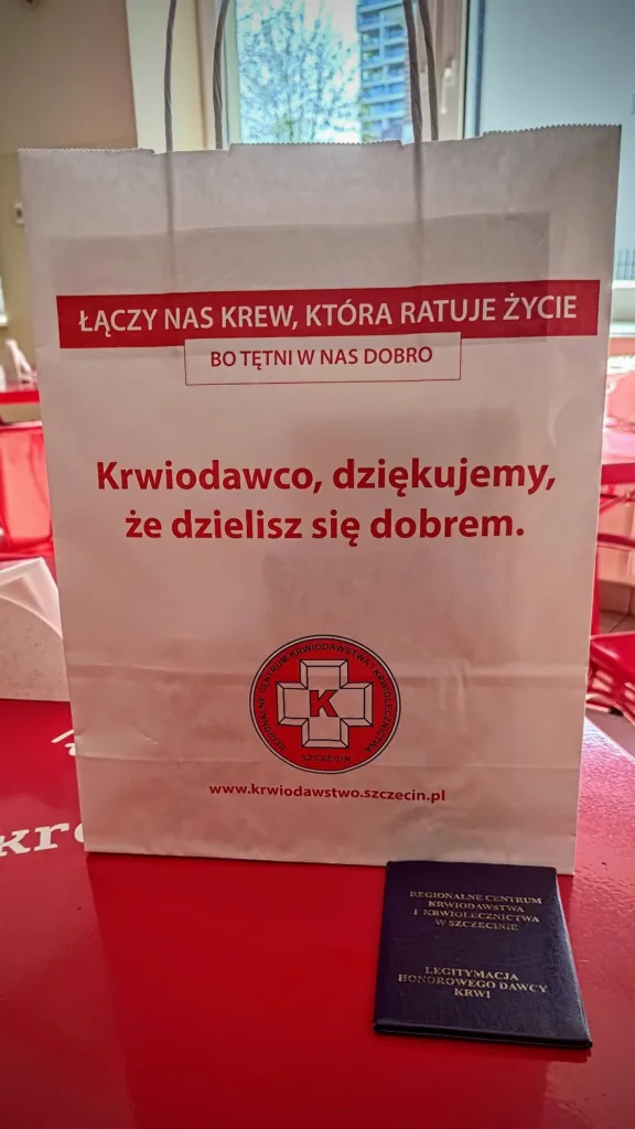 Biała torba z czerwonymi napisami i logo Regionalnego Centrum Krwiodawstwa i Krwiolecznictwa w Szczecinie, obok leży granatowa legitymacja honorowego dawcy krwi.