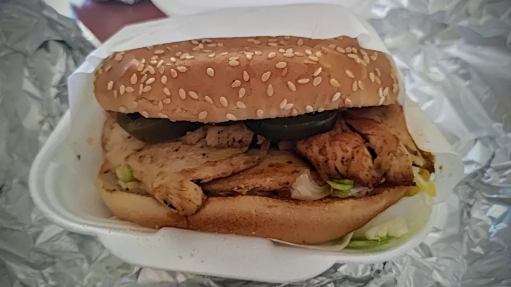 Kanapka z grillowanym kurczakiem, sałatą i plasterkami jalapeno w bułce sezamowej, podana w białym styropianowym pudełku.