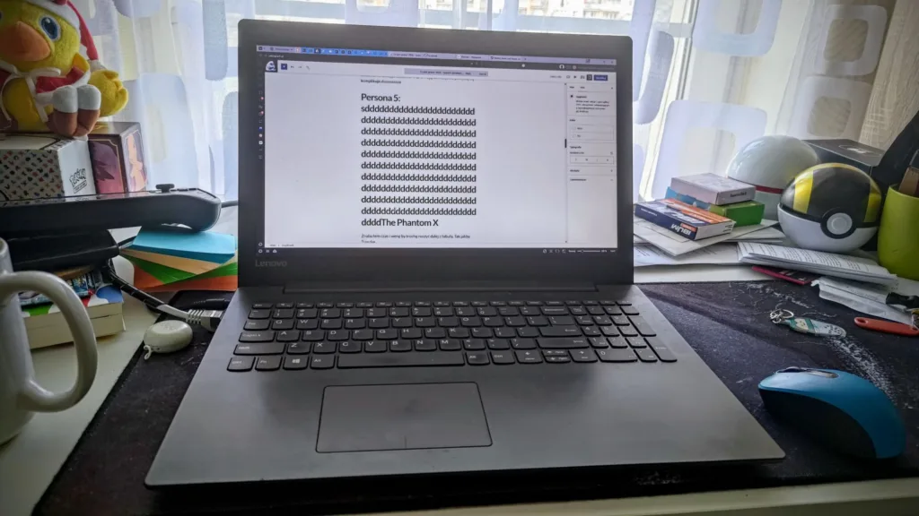 Laptop z otwartym dokumentem tekstowym na ekranie, na biurku obok kubka, pluszowej zabawki i przedmiotów związanych z grami.