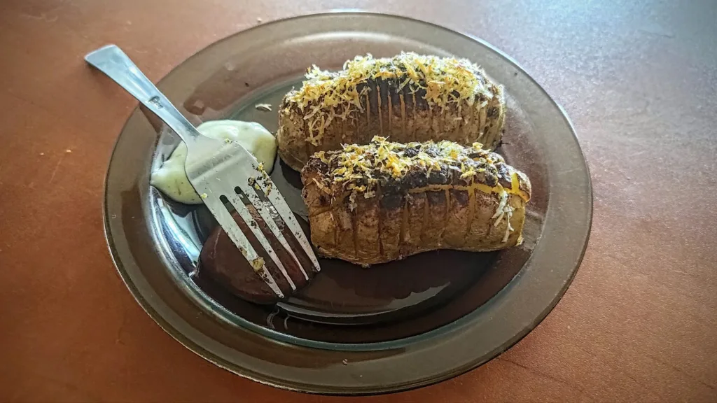 Dwa pieczone ziemniaki Hasselback posypane serem i ziołami, podane na ciemnym talerzu z widelcem i sosem.