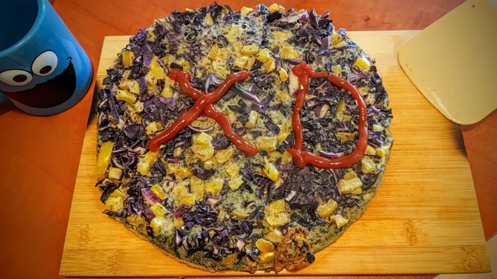 Okrągły placek z warzywami, ozdobiony keczupem w kształcie napisu "XD". Obok znajduje się niebieski kubek z postacią z kreskówki.