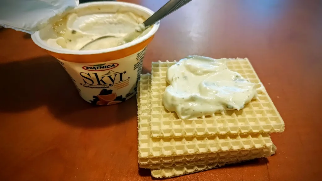 Kubek jogurtu typu islandzkiego Skyr o smaku pomarańczy z czekoladą, obok stos wafli z nałożonym kremowym jogurtem.