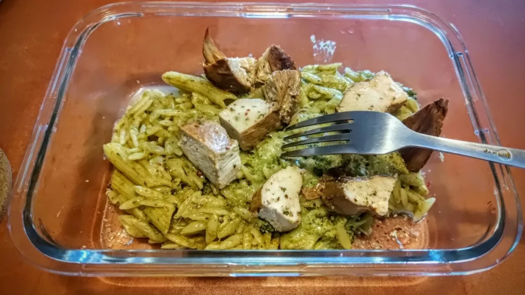 Makaron orzo z sosem pesto i kawałkami pieczonego kurczaka w szklanym pojemniku, widelcem w daniu.