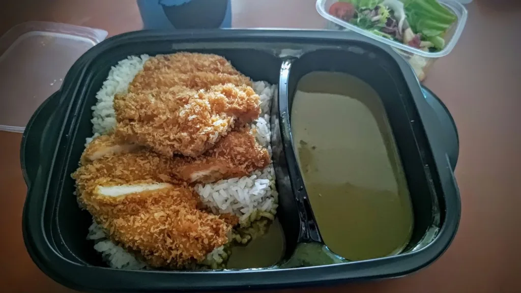 Kotlet panierowany z kurczaka podany z ryżem i sosem curry w czarnym pojemniku na wynos.
