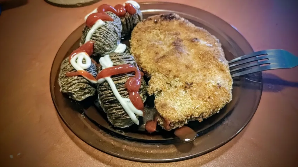 Talerz z pieczonymi ziemniakami Hasselback polanymi keczupem i majonezem oraz panierowanym kotletem.