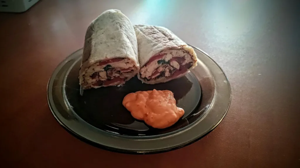 Dwa kawałki wrapa z kurczakiem, pomidorami i zielonymi liśćmi, podane z pomarańczowym sosem na czarnym talerzu.