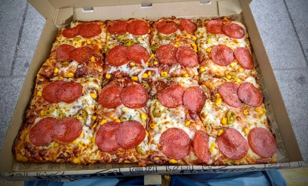 Duża, prostokątna pizza z pepperoni, kukurydzą i zielonymi papryczkami, pokrojona na kwadratowe kawałki, w otwartym kartonowym pudełku.