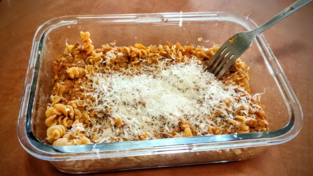 Makaron fusilli z sosem mięsnym i startym serem w szklanym naczyniu, widelcem w potrawie.