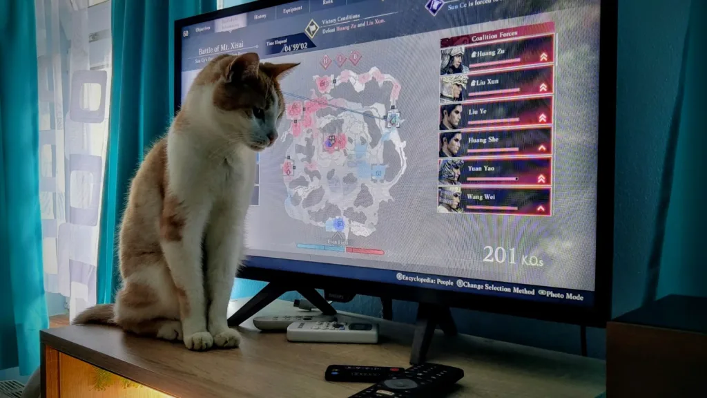 Kot siedzący na biurku przed monitorem, na którym wyświetlana jest mapa z gry komputerowej Dynasty Warriors.