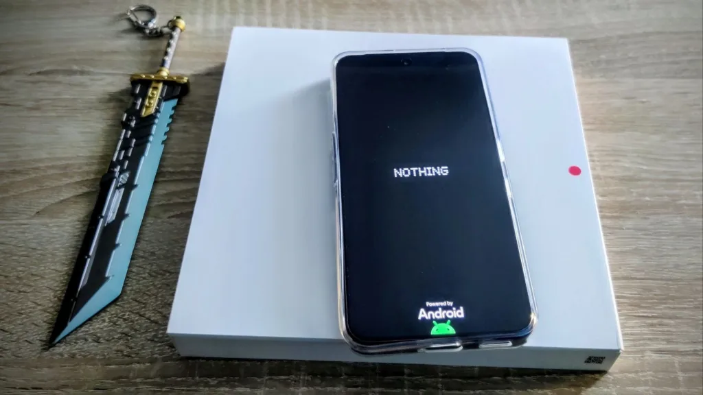Smartfon marki Nothing na białym pudełku, obok breloczek w kształcie miecza.