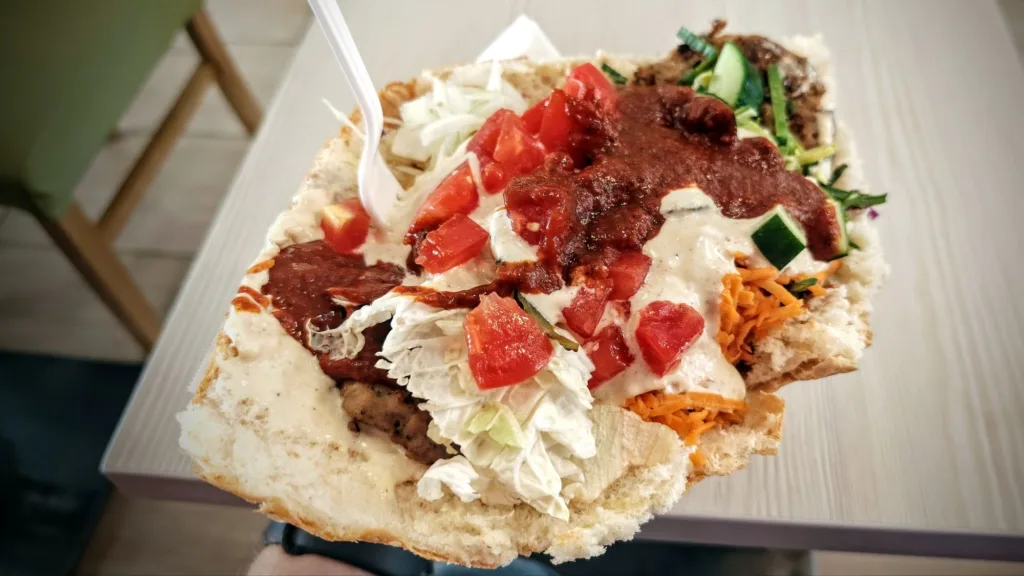 Duży hamburger w bułce od kebaba, z warzywami i sosami, trzymany w ręce z widocznym widelcem.