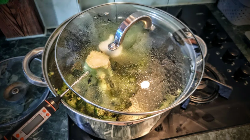 Gotujący się brokuł w garnku pod szklaną pokrywą, z widocznym termometrem kuchennym