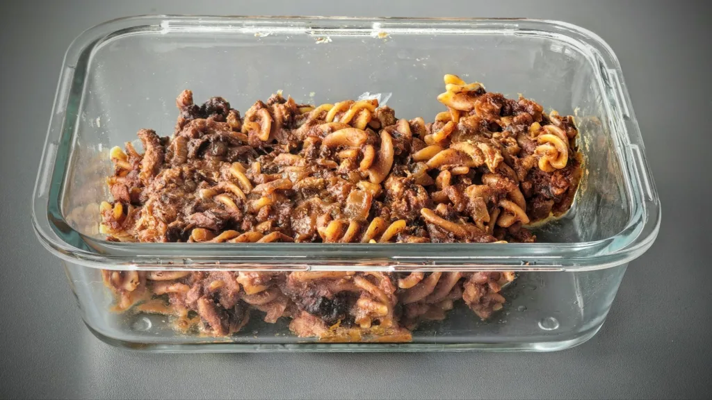 Makaron w kształcie ślimaków wymieszany z gulaszem, podany w szklanym pojemniku na ciemnym tle.