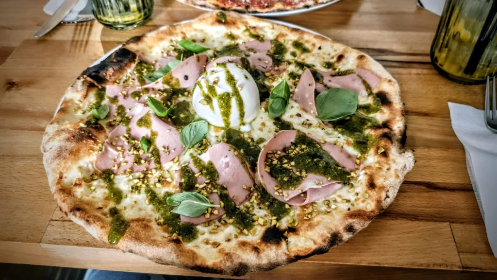 Pizza z plasterkami mortadeli, zielonym sosem i pistacjami, z kulką burraty na środku.