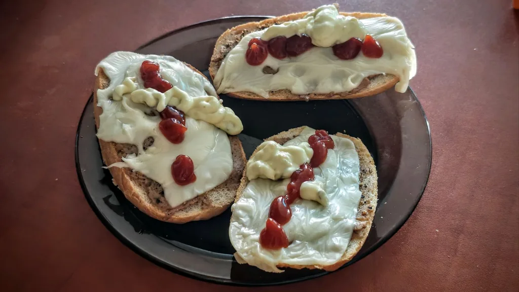 Trzy kromki chleba na czarnym talerzu, posmarowane serem, z dodatkiem ketchupu.