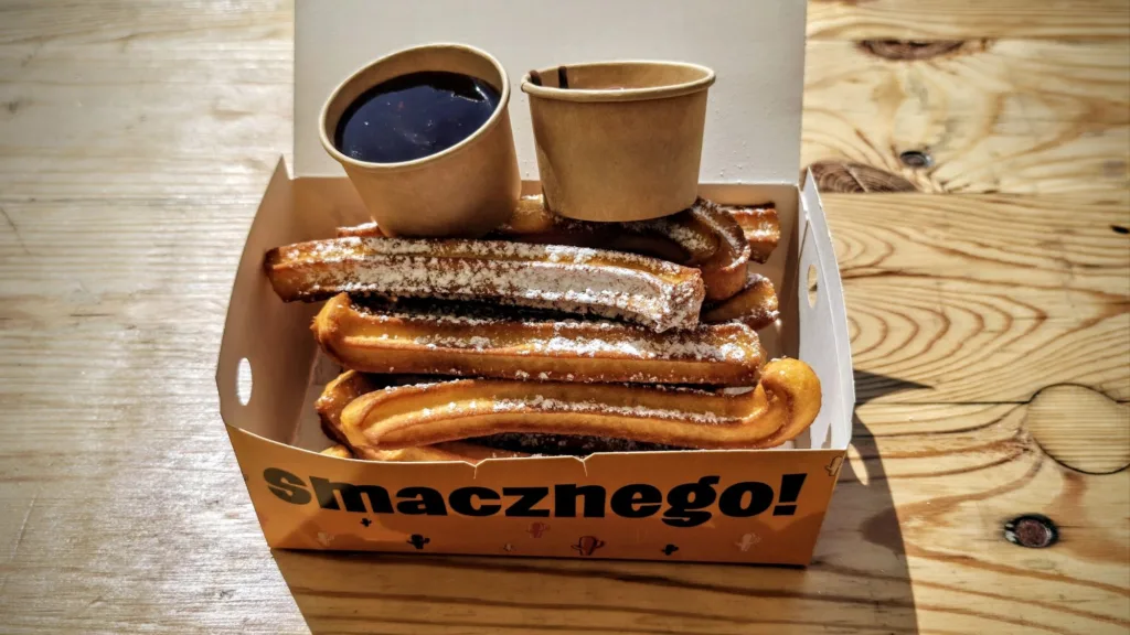 Stos chrupiących churrosów posypanych cukrem pudrem, podawanych w kartonowym pudełku z dwoma kubeczkami sosu do maczania na drewnianym tle.