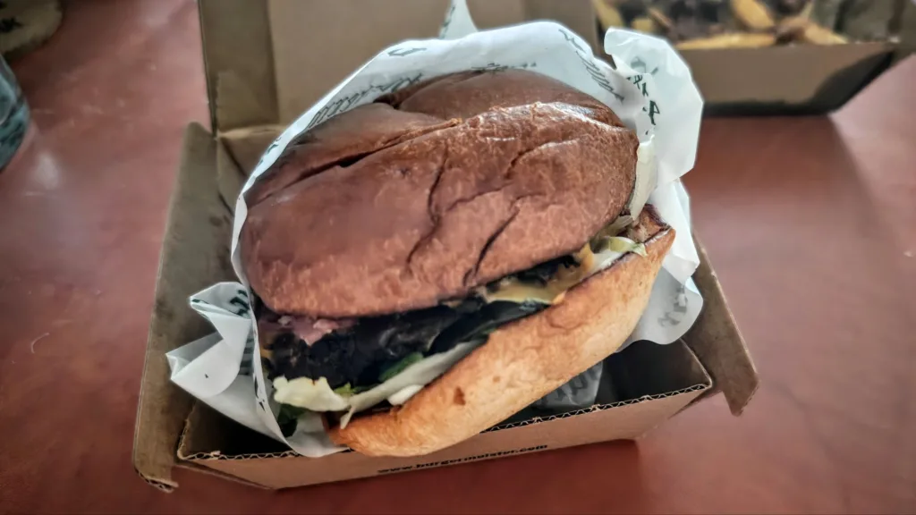Duży burger w bułce sezamowej z sałatą, mięsem i sosem, zawinięty w białą papierową serwetkę, leżący w brązowym kartonowym opakowaniu.