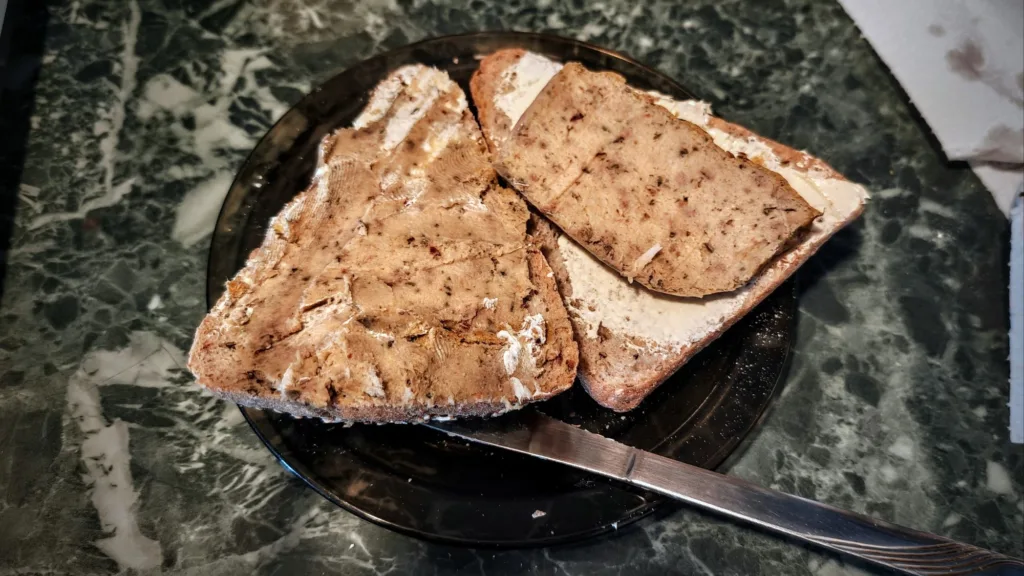 Dwa kanapki z chlebem posmarowane czymś jasnym, posypane przyprawami z pasztetem na wierzchu, leżące na czarnym talerzu.