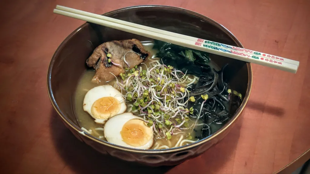 Miseczka ramen z makaronem, mięsem, połówkami jajka, kiełkami i ciemnymi glonami, przykryta pałeczkami do jedzenia.