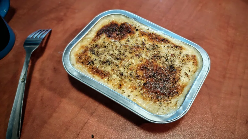 Zapiekanka w aluminiowej tackce, z przypieczonym serem i ziołami, obok leży widelec.