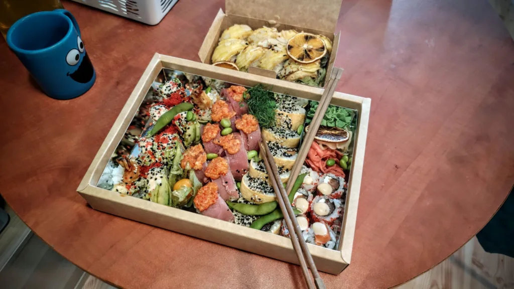 Dwa pudełka z sushi i rolkami na drewnianym stole, obok niebieskiego kubka.