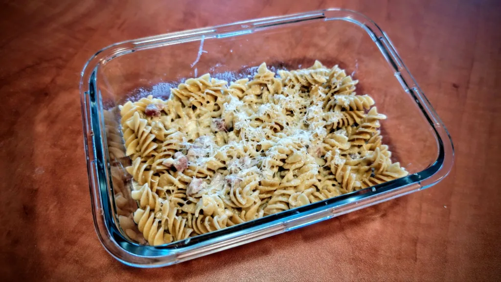 Makaron fusilli w sosie śmietanowo-serowym z kawałkami boczku, posypany startym parmezanem, podany w szklanym naczyniu.