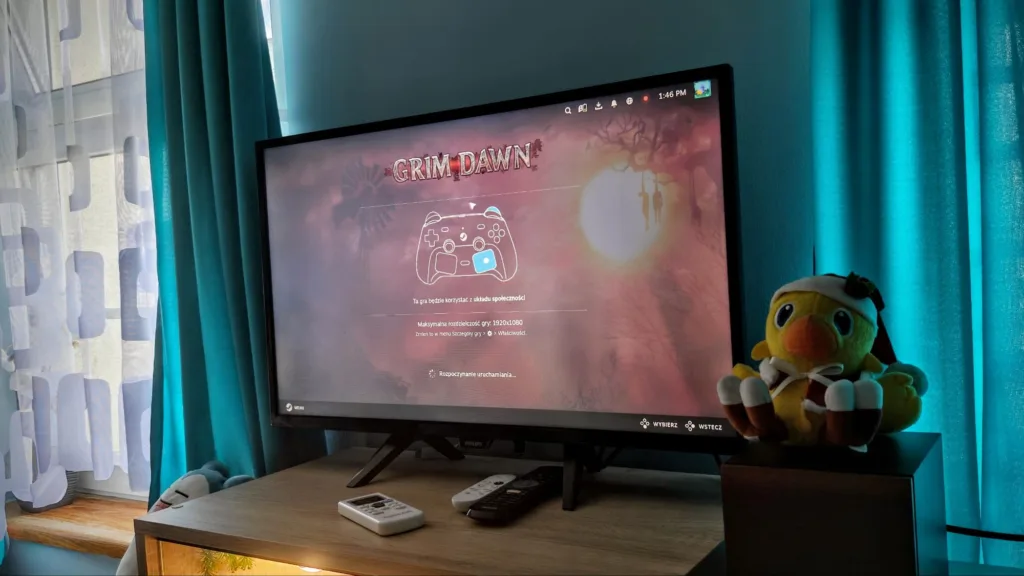 Włączony telewizor pokazujący ekran startowy gry Grim Dawn z kontrolerem widocznym na ekranie. Po prawej stronie stoi pluszowa zabawka w kształcie żółtego ptaka z czerwoną czapką.