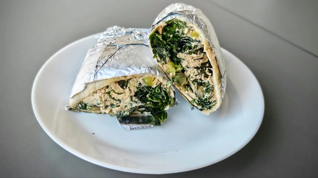 Dwa kawałki wrapa zawiniętego w folię aluminiową, wypełnione zielonymi liśćmi, białą masą i kawałkami mięsa.