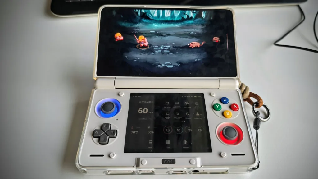 Przenośna konsola do gier w otwartym stanie, z widocznym ekranem wyświetlającym scenę z gry fantasy z postaciami i potworami, oraz panelem sterowania z przyciskami i joystickami.