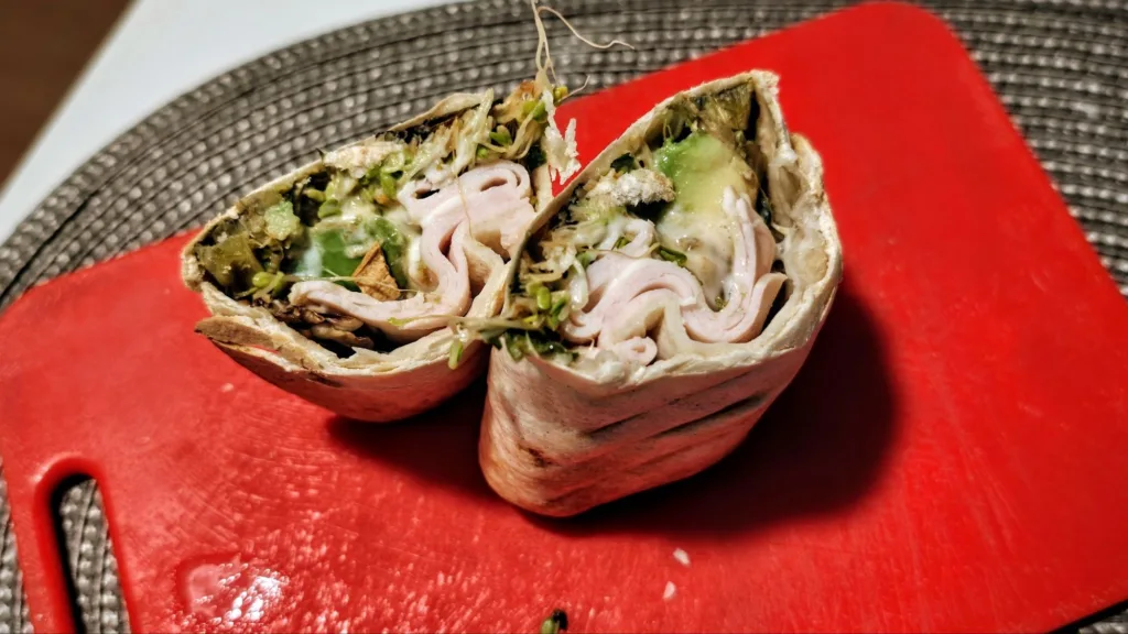 Przekrojona na pół tortilla z wędliną, awokado i kiełkami, leżąca na czerwonej desce do krojenia.