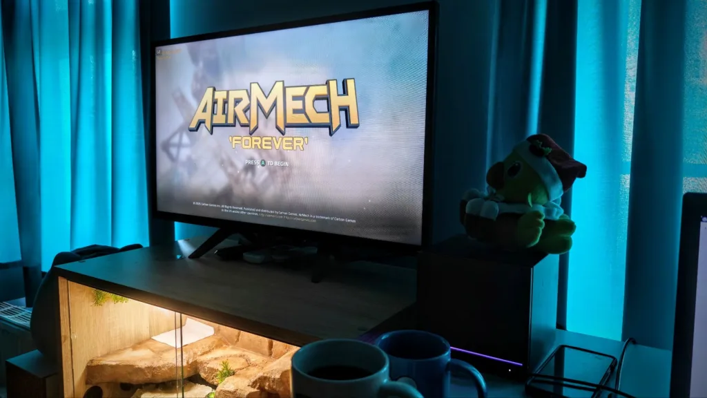 Ekran telewizora z tytułem gry AirMech w pokoju oświetlonym na niebiesko, obok maskotki i kubków na biurku.