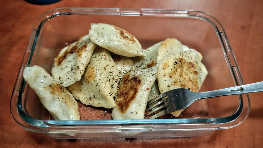 Usmażone pierogi posypane pieprzem i ziołami w szklanym pojemniku z widocznym widelcem.