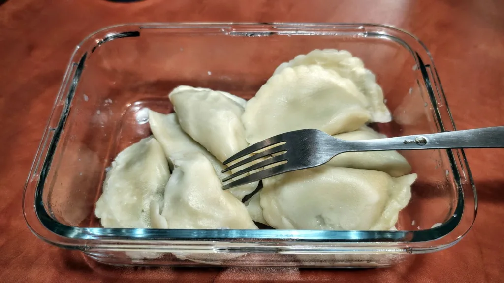 Kilka ugotowanych pierogów w szklanym naczyniu na brązowym blacie, na jednym z nich leży metalowy widelca.