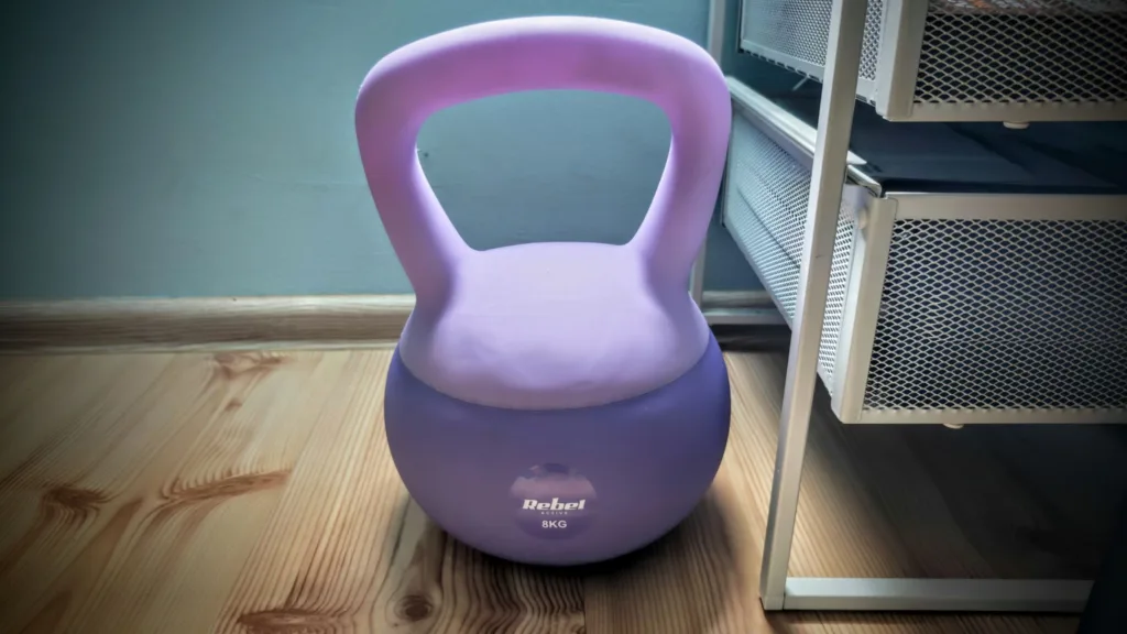 Fioletowy odważnik kettlebell Rebel Active 8 kg stoi na drewnianej podłodze obok białego regału.